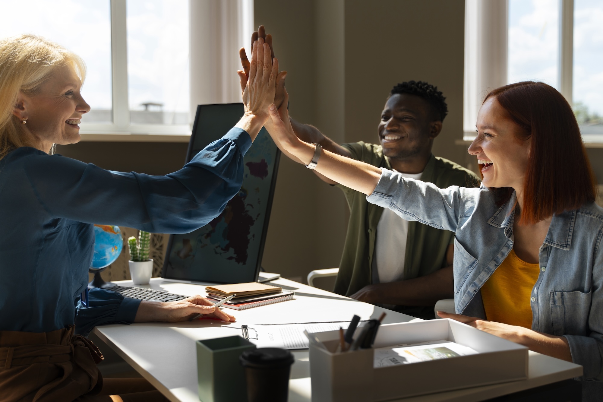 Three colleagues in a bright office high-fiving each other with smiles on their faces. The image depicts a sense of teamwork, positivity, and success, illustrating the concept of employee resilience. This captures the essence of a resilient workforce, where individuals support each other, adapt to challenges, and maintain a positive and productive work environment.